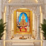 Swaminarayan Maharaj in elaborate gold and red devotional attire, framed by ornate temple pillars and flower offerings.